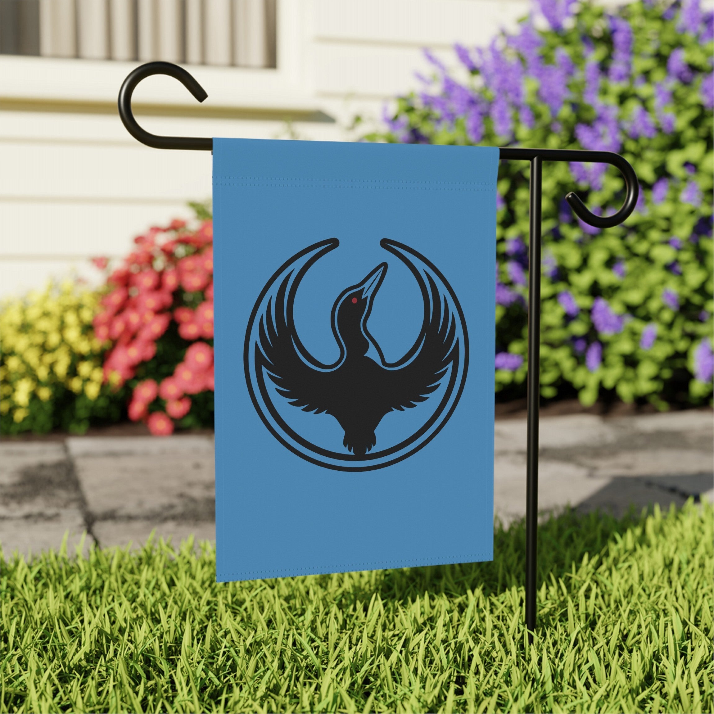 Minnesota Rebel Alliance Garden Flag featuring a stylized loon emblem on light-blue fabric, mounted on a black stand in a lawn, symbolizing resistance and immigrant rights advocacy. Size: 12 x 18.