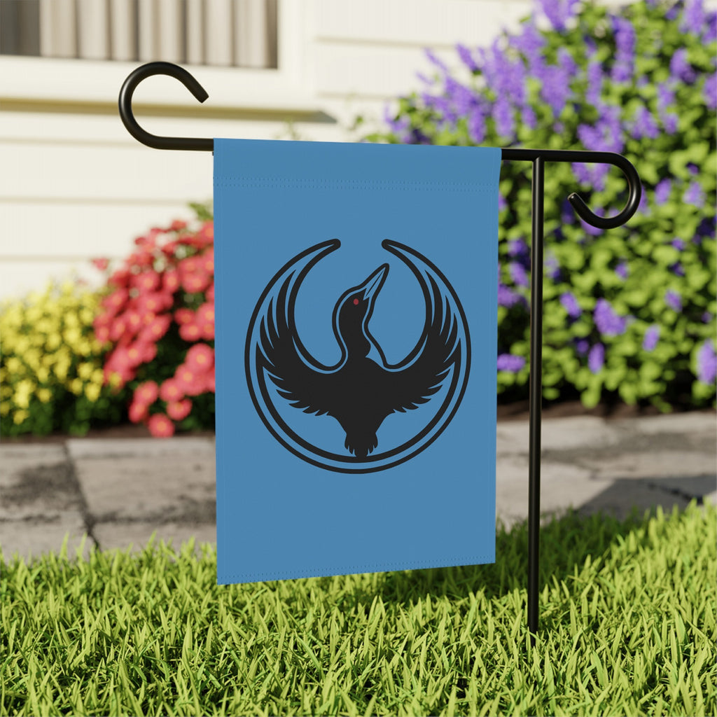 Minnesota Rebel Alliance Garden Flag featuring a stylized loon emblem on light-blue fabric, mounted on a black stand in a lawn, symbolizing resistance and immigrant rights advocacy. Size: 12 x 18.
