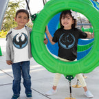 Two children wearing Minnesota Rebel Alliance T-shirts with a loon emblem play on a green playground ring, smiling brightly. The design reflects bold, non-neutral activism through fashion.
