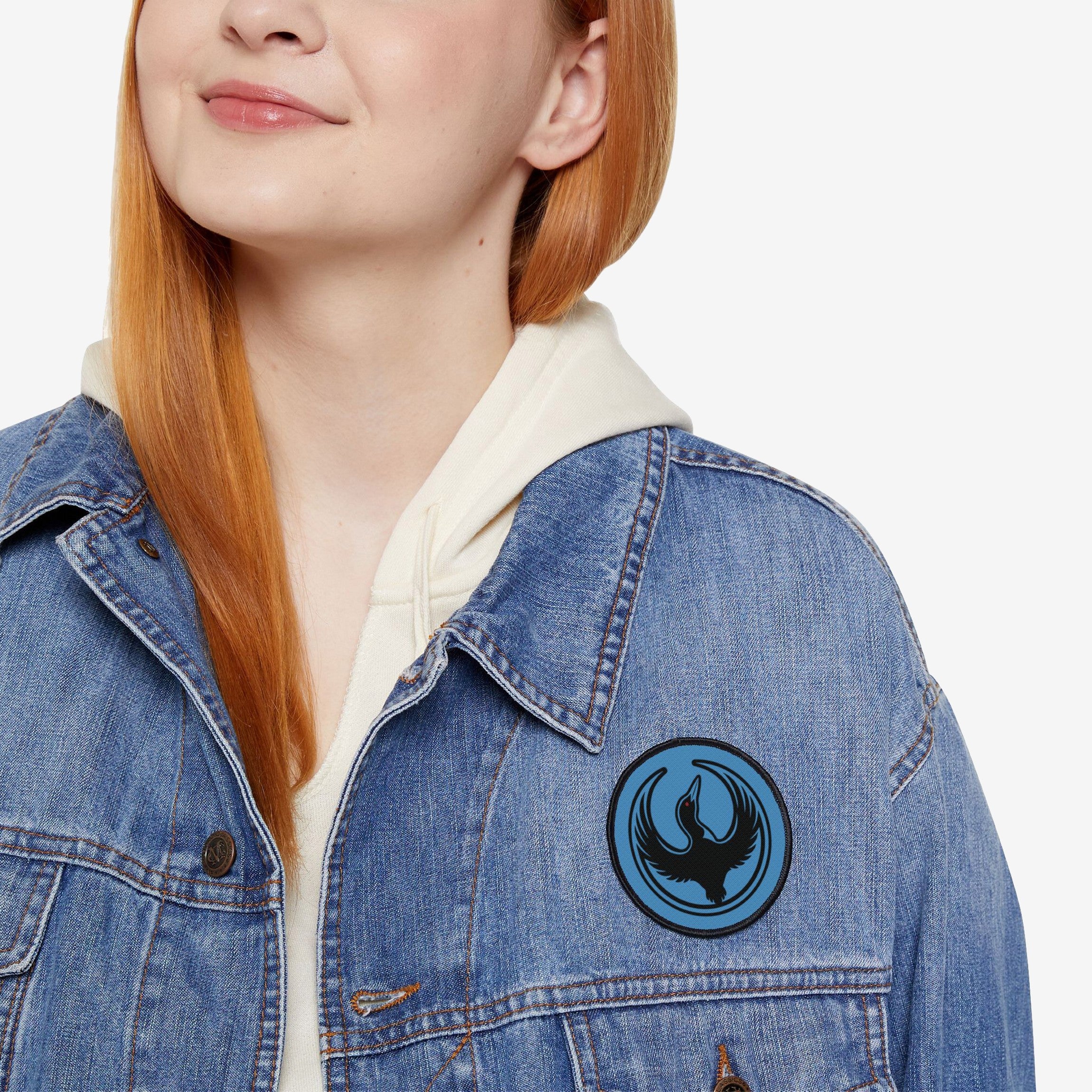 Red-haired person wearing a light-wash denim jacket with a Minnesota Rebel Alliance Patch featuring a stylized black loon on a blue circle, symbolizing activism and equality, over a cream hoodie.