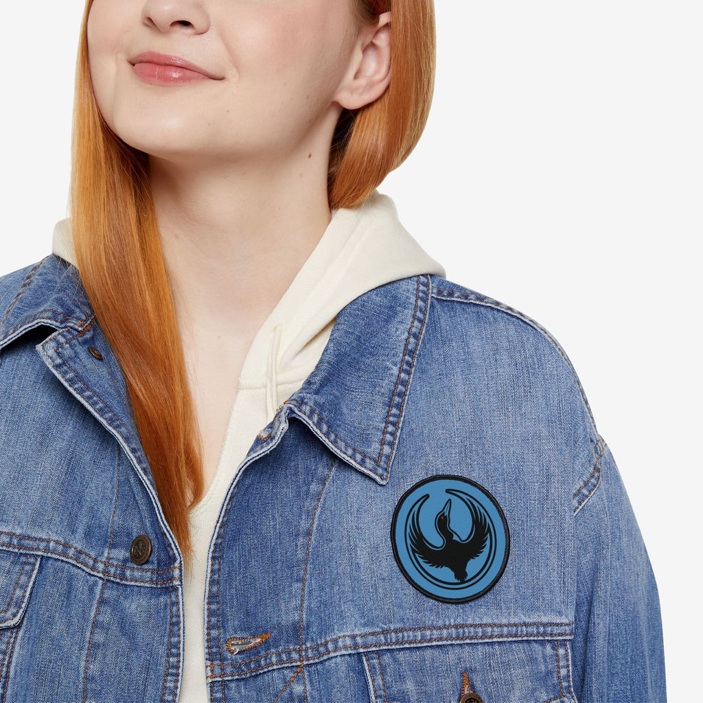 Red-haired person wearing a light-wash denim jacket with a Minnesota Rebel Alliance Patch featuring a stylized black loon on a blue circle, symbolizing activism and equality, over a cream hoodie.