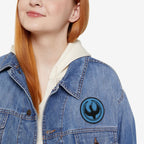 Red-haired person wearing a light-wash denim jacket with a Minnesota Rebel Alliance Patch featuring a stylized black loon on a blue circle, symbolizing activism and equality, over a cream hoodie.