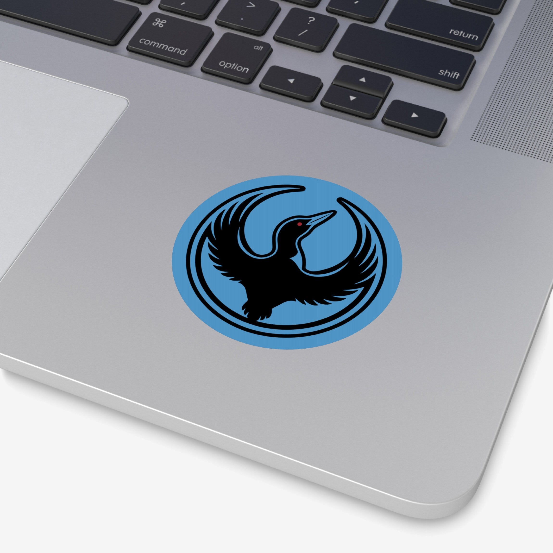Close-up of a silver laptop featuring the Minnesota Rebel Alliance Sticker, a round blue design with a black loon logo, symbolizing resistance against oppression and support for immigrant rights.