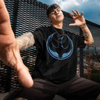 Young person wearing a black Minnesota Rebel Alliance T-shirt with a bold loon graphic, crouching in front of a cityscape. Their arms are tattooed, adding an urban, fashion-forward edge.