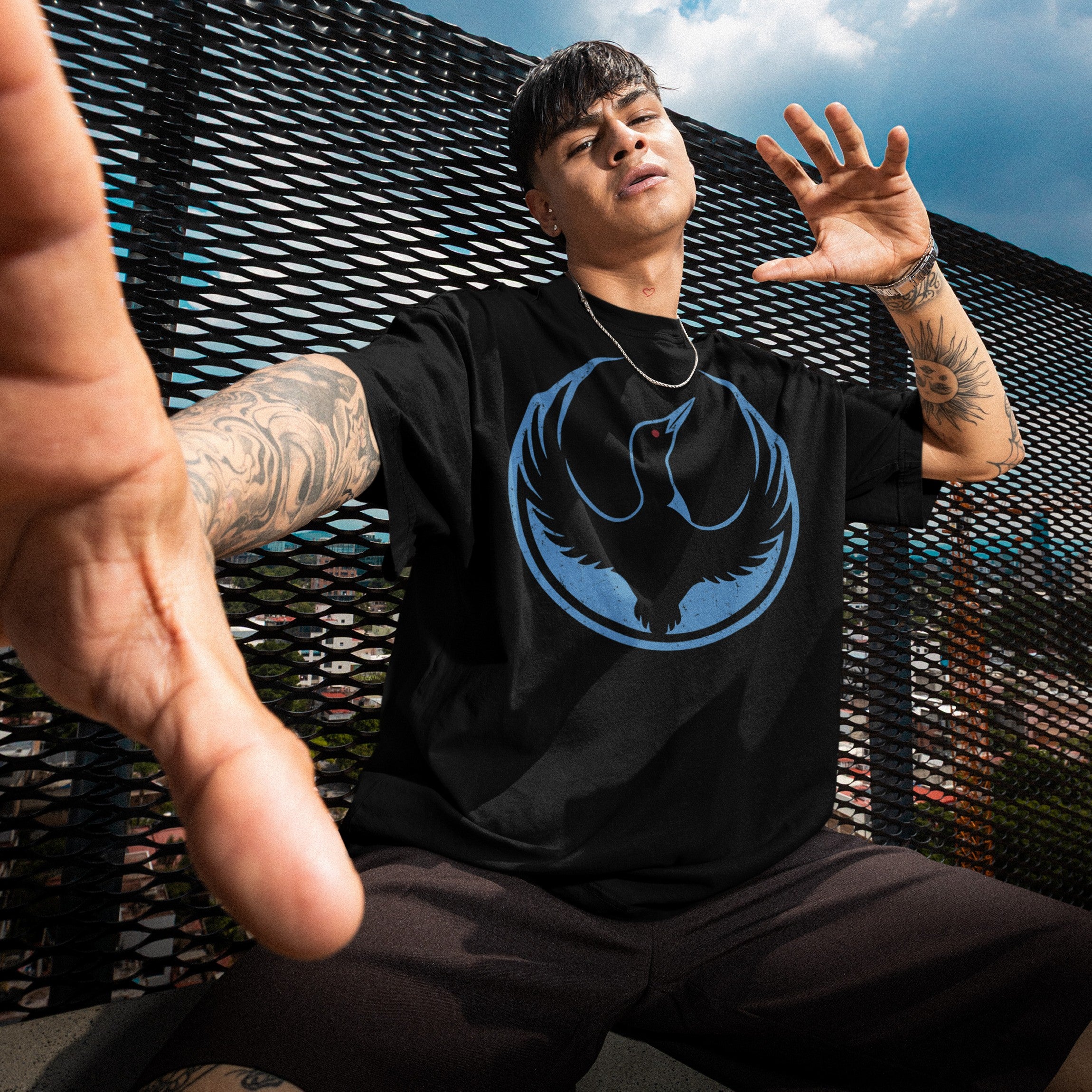 Young person wearing a black Minnesota Rebel Alliance T-shirt with a bold loon graphic, crouching in front of a cityscape. Their arms are tattooed, adding an urban, fashion-forward edge.