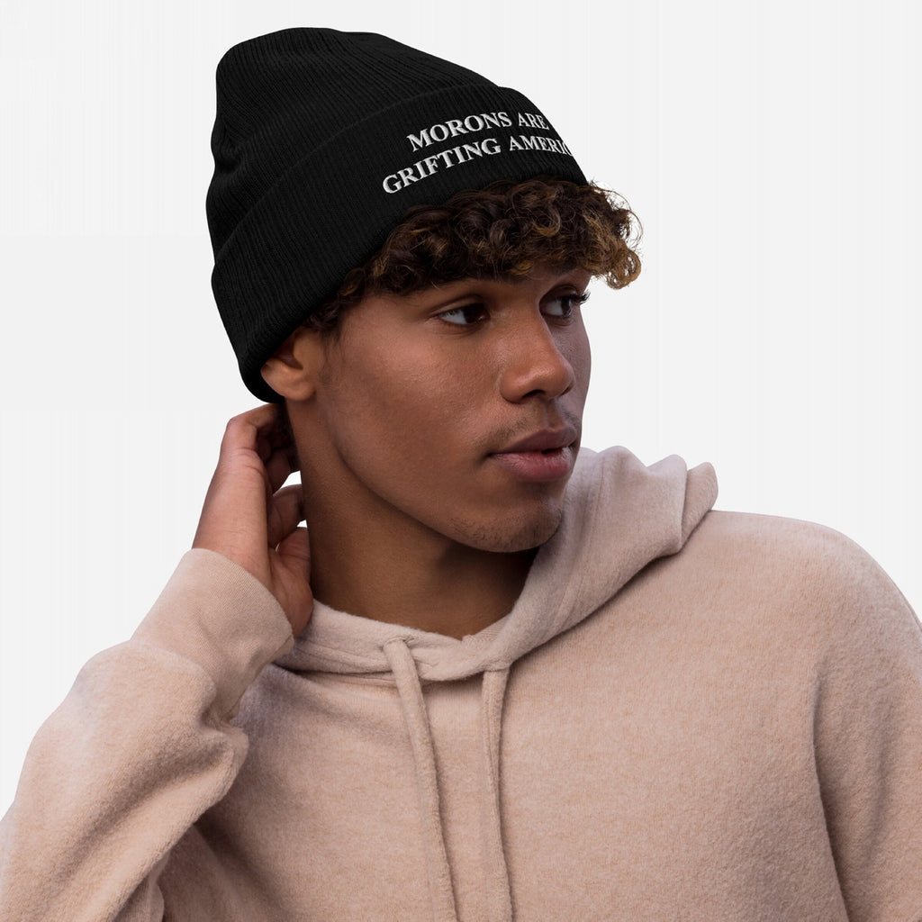 A lifestyle image of a person wearing the black anti-MAGA beanie embroidered with "MORONS ARE GRIFTING AMERICA," styled casually with a light beige hoodie, showcasing the hat’s fit and bold message in everyday wear.