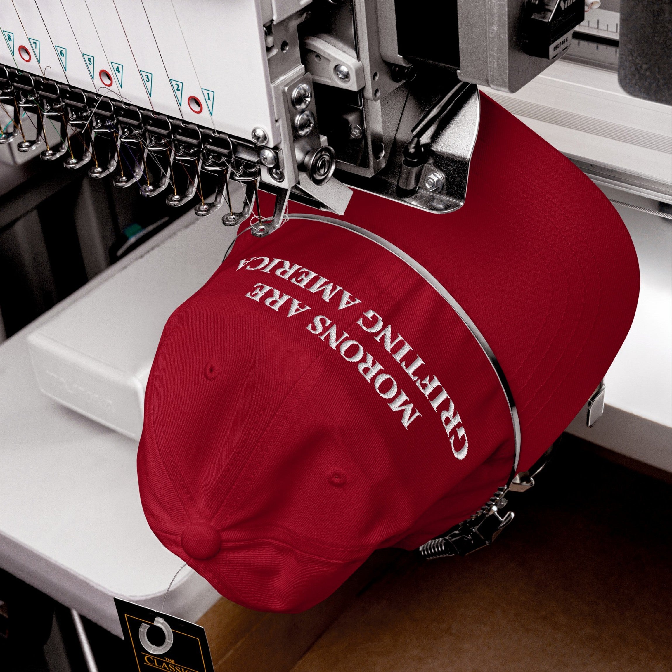 Machine embroidery in progress stitching "Morons Are Grifting America" onto a red hat — a high-quality production of a bold anti-MAGA design.