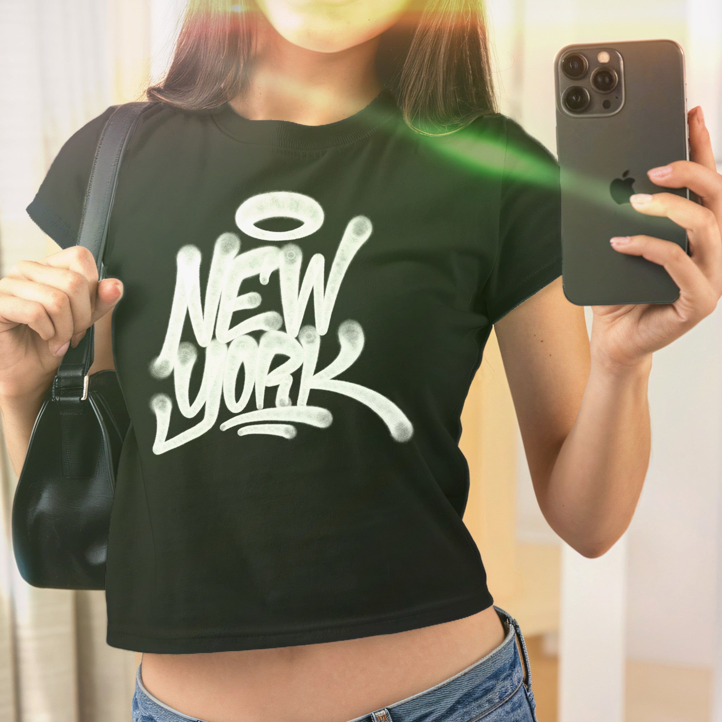 Person in mirror selfie wearing the New York Handstyle Women's Baby Tee, featuring a white graffiti-style logo with a halo on a black crop-style T-shirt, paired with blue denim jeans.