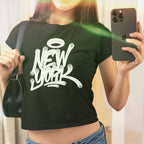Person in mirror selfie wearing the New York Handstyle Women's Baby Tee, featuring a white graffiti-style logo with a halo on a black crop-style T-shirt, paired with blue denim jeans.