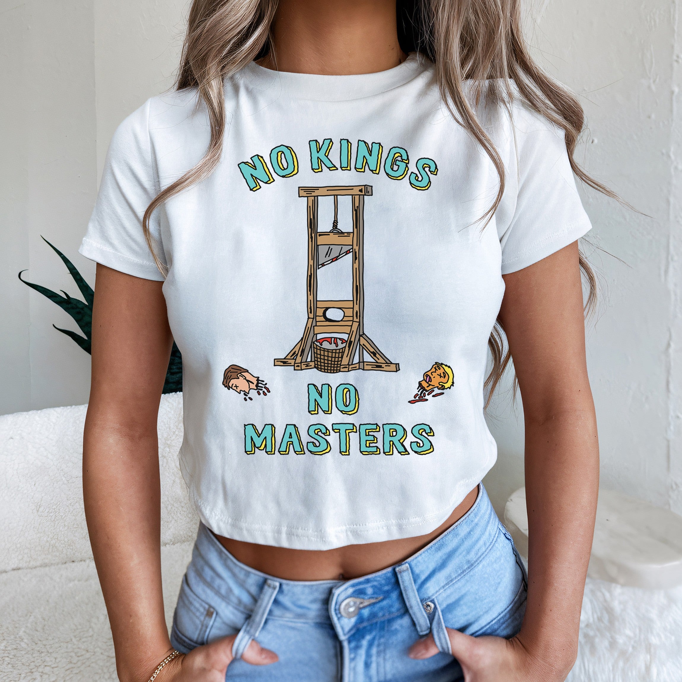 No Kings No Masters Women's Baby Tee featuring a guillotine with two cartoon heads and bold anti-authoritarian text. The cropped design embodies a rebellious spirit with a gritty, hand-drawn style.