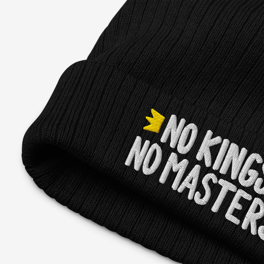 No Kings No Masters Hat features a black rib-knit cuffed design with white embroidered text and a yellow crown icon, embodying rebellious spirit and defiance against oppressive systems.