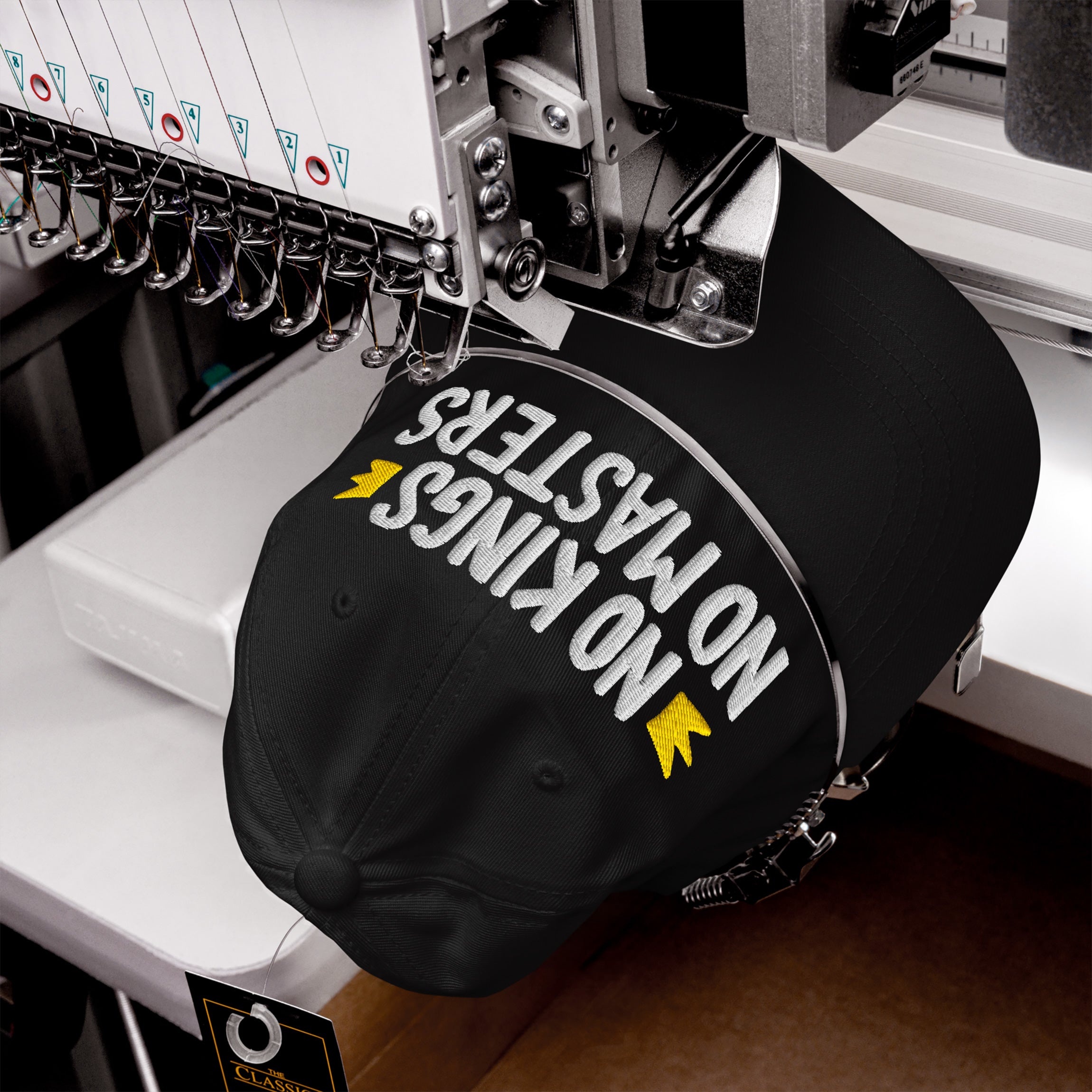 No Kings No Masters Hat in an embroidery machine, featuring white and yellow stitching. The cap's message embodies defiance and individuality, aligning with a bold, anti-establishment ethos.