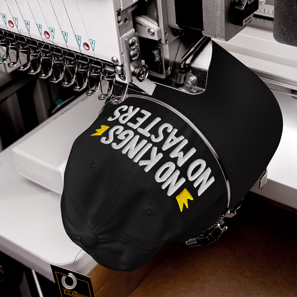 No Kings No Masters Hat in an embroidery machine, featuring white and yellow stitching. The cap's message embodies defiance and individuality, aligning with a bold, anti-establishment ethos.