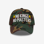The No Kings No Masters Hat features a camouflage design with bold white embroidery and yellow crown accents, embodying rebellious spirit against authority, aligning with an ethos of equality and anti-fascism.