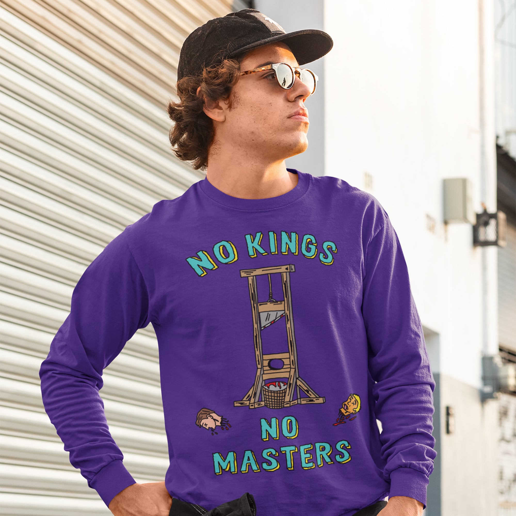 Young man outdoors wearing the No Kings No Masters Shirt featuring a guillotine illustration with severed heads, symbolizing anti-authoritarian defiance. The shirt advocates resistance against fascism and supports equality.