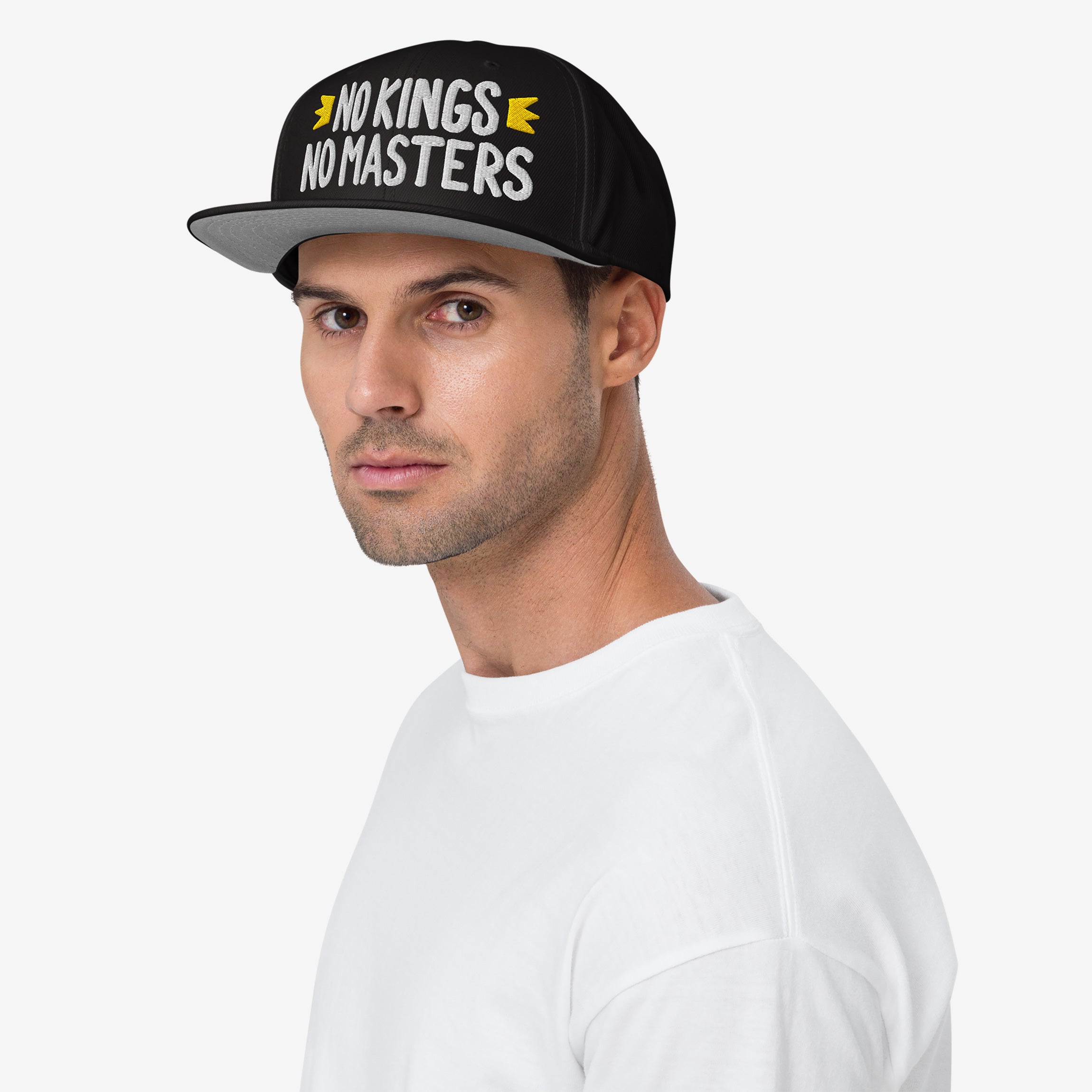 Young man in a white T-shirt wears the No Kings No Masters Hat, a black snapback with white embroidery challenging authority, featuring yellow arrows. Symbolizes rebellion and individuality against oppressive systems.