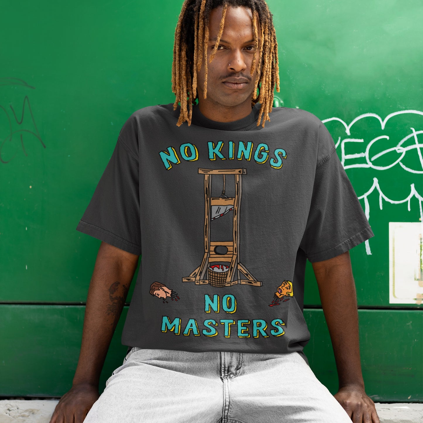 Young man wearing the No Kings No Masters T-shirt with a graphic guillotine and severed heads, embodying rebellion. Casual style with dreadlocks and a nose ring, against a graffiti-tagged wall.