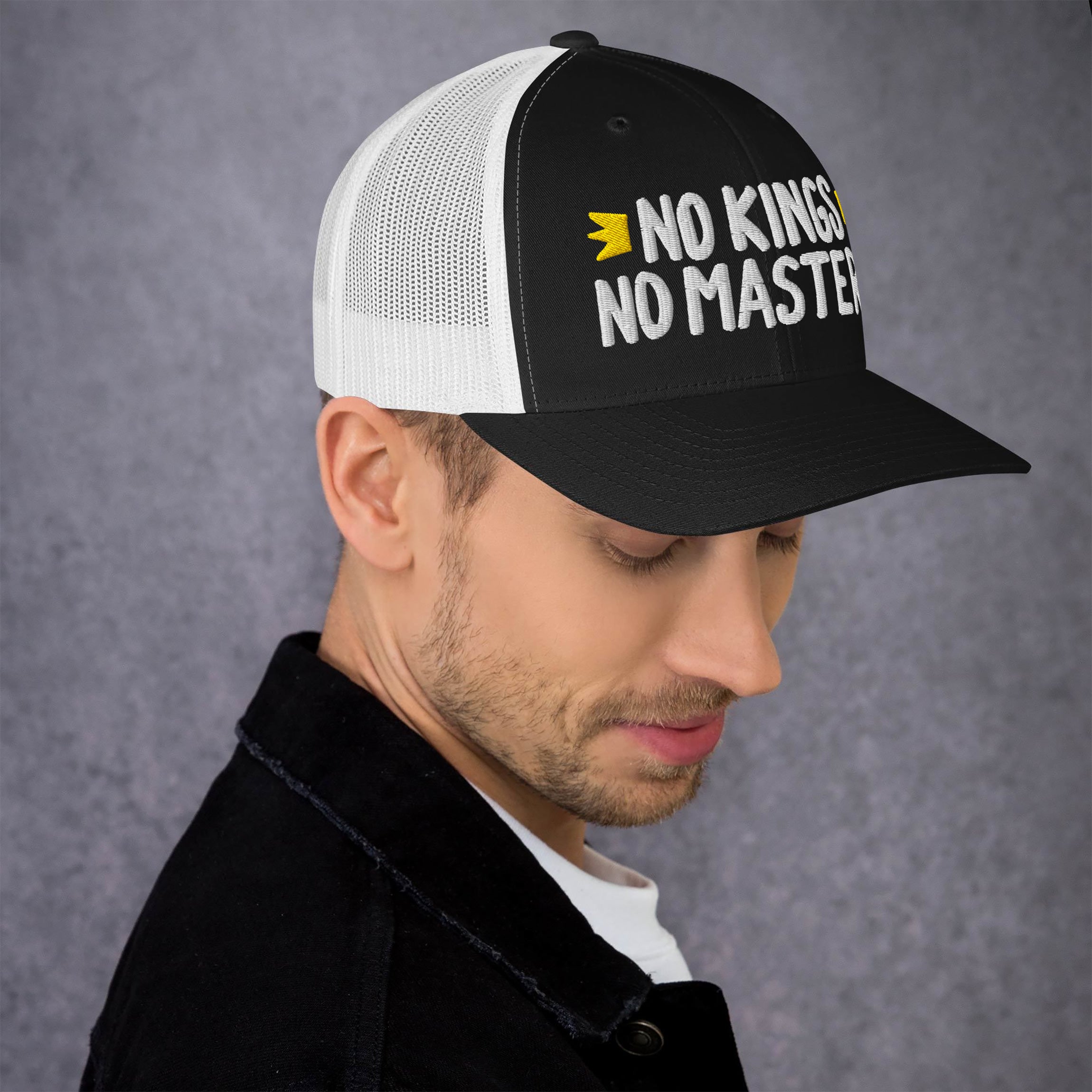 Young man in profile wearing the No Kings No Masters Hat, a black-and-white mesh trucker cap with bold white lettering and a yellow crown icon, symbolizing a stand against oppressive systems.