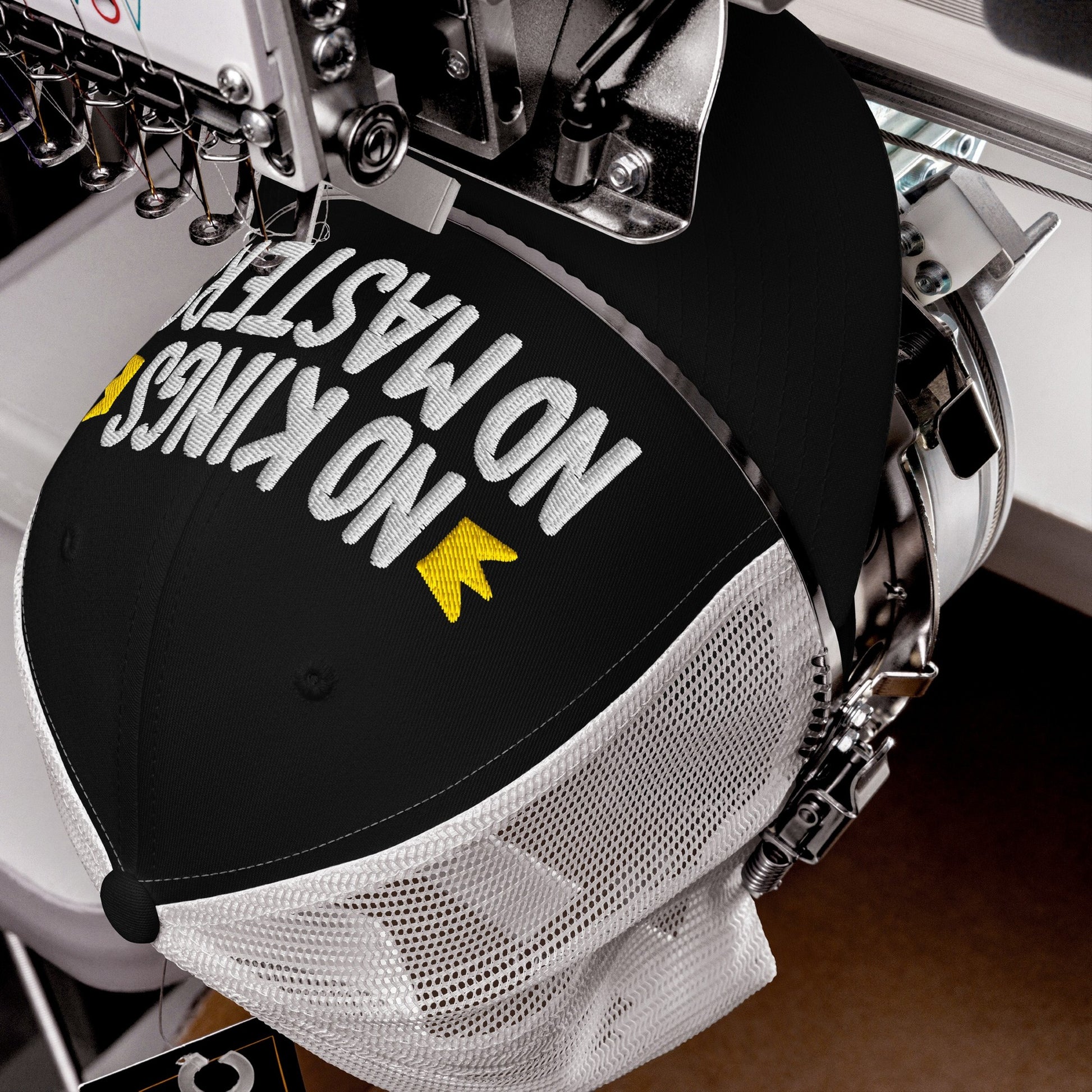 The No Kings No Masters Hat in an embroidery machine, stitching bold white letters with yellow accents on a black-and-white mesh trucker cap, embodying rebellion and modern resistance.