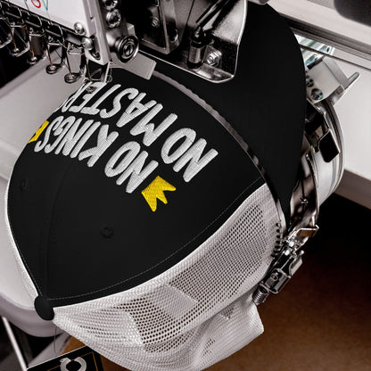 The No Kings No Masters Hat in an embroidery machine, stitching bold white letters with yellow accents on a black-and-white mesh trucker cap, embodying rebellion and modern resistance.