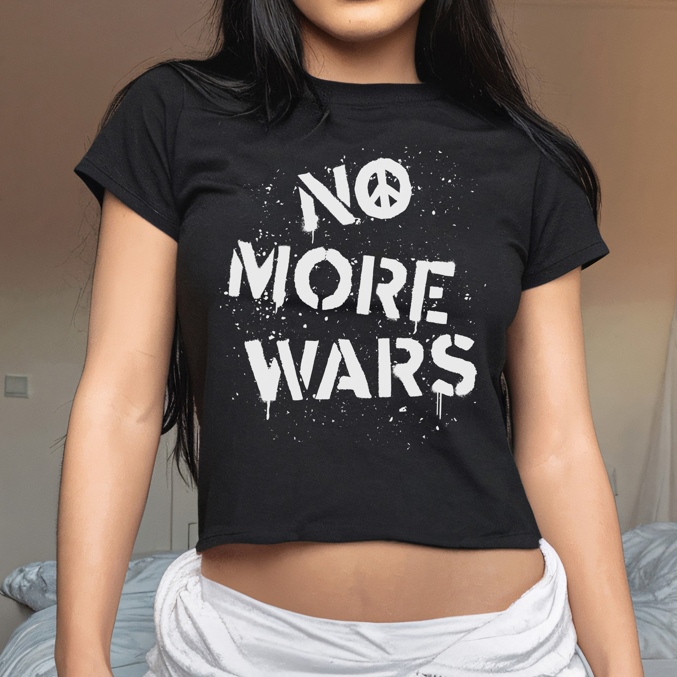 No More Wars Women's Baby Tee featuring a bold stencil-style slogan with a peace symbol, worn by a woman indoors, symbolizing protest and solidarity against global conflicts.