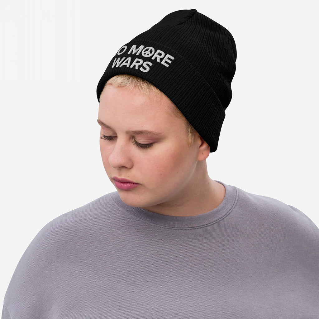 A person wears the No More Wars Hat, featuring the slogan and peace symbol embroidery on a black beanie, embodying a message of peace. They pair it with a light gray sweatshirt.
