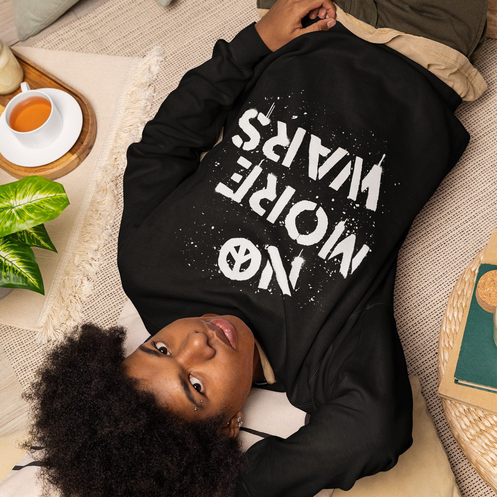 Person reclining on a woven mat wearing a black No More Wars sweatshirt. Nearby, a wooden tray with tea and a plant, and a wicker table with books, reflect a peaceful, contemplative scene.