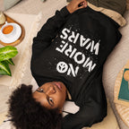 Person reclining on a woven mat wearing a black No More Wars sweatshirt. Nearby, a wooden tray with tea and a plant, and a wicker table with books, reflect a peaceful, contemplative scene.