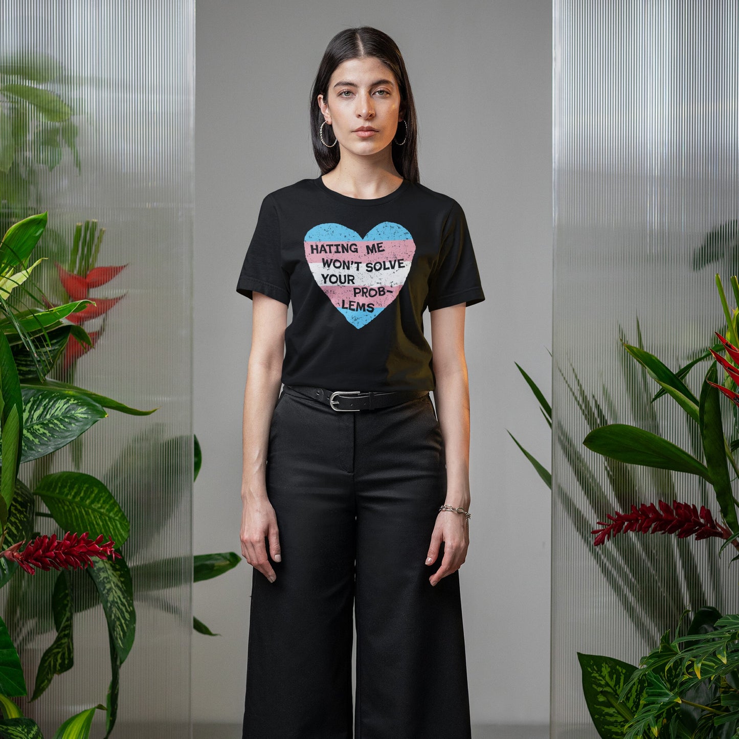 Woman in black No Trans Hate T-shirt with trans pride heart graphic, stating Hating Me Won’t Solve Your Problems, stands confidently between greenery, embodying strength and visibility.