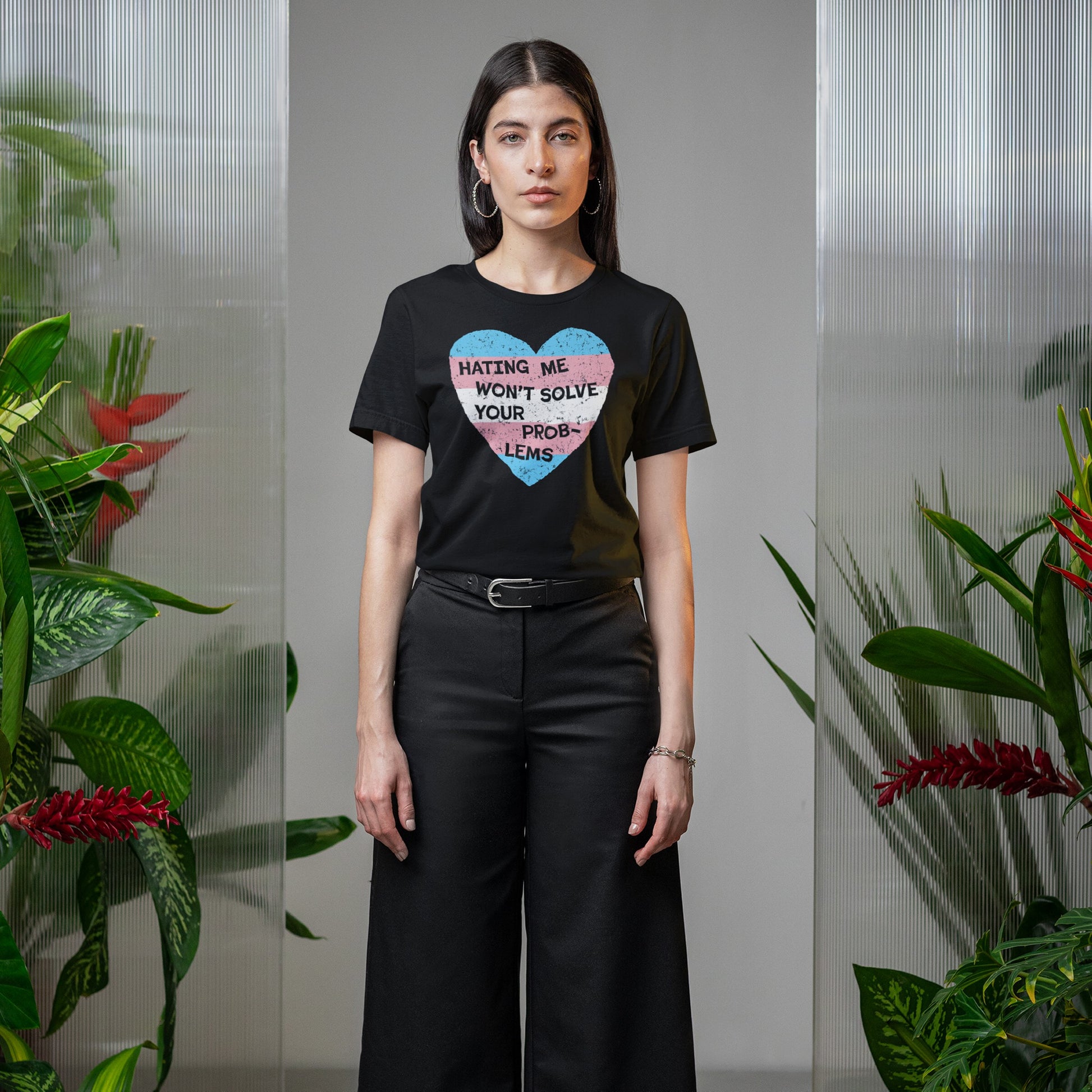 Woman in black No Trans Hate T-shirt with trans pride heart graphic, stating Hating Me Won’t Solve Your Problems, stands confidently between greenery, embodying strength and visibility.