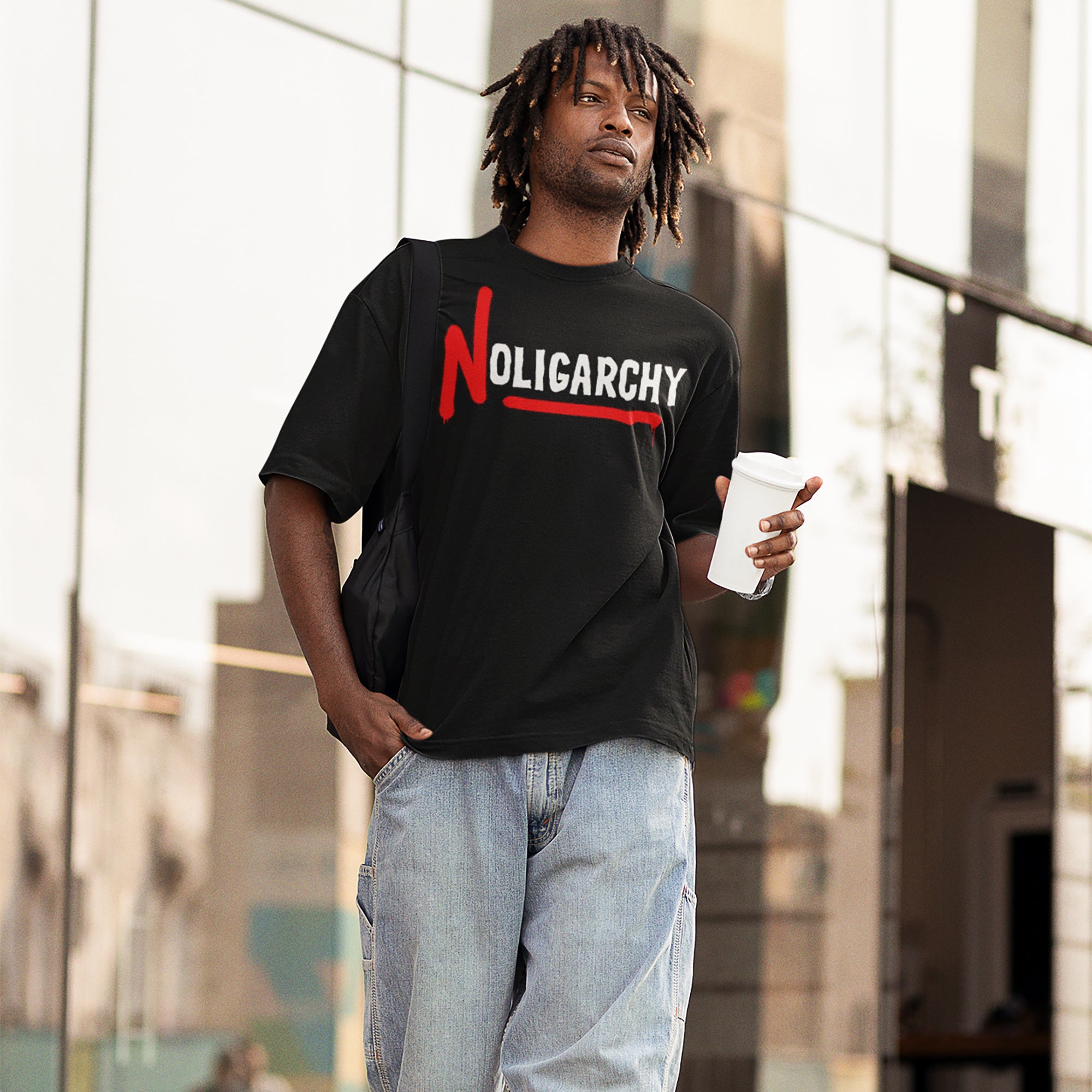 Man wears a No Oligarchy Shirt in an urban setting, featuring bold graffiti-style text. He carries a black bag and a coffee cup, embodying rebellion and activism.