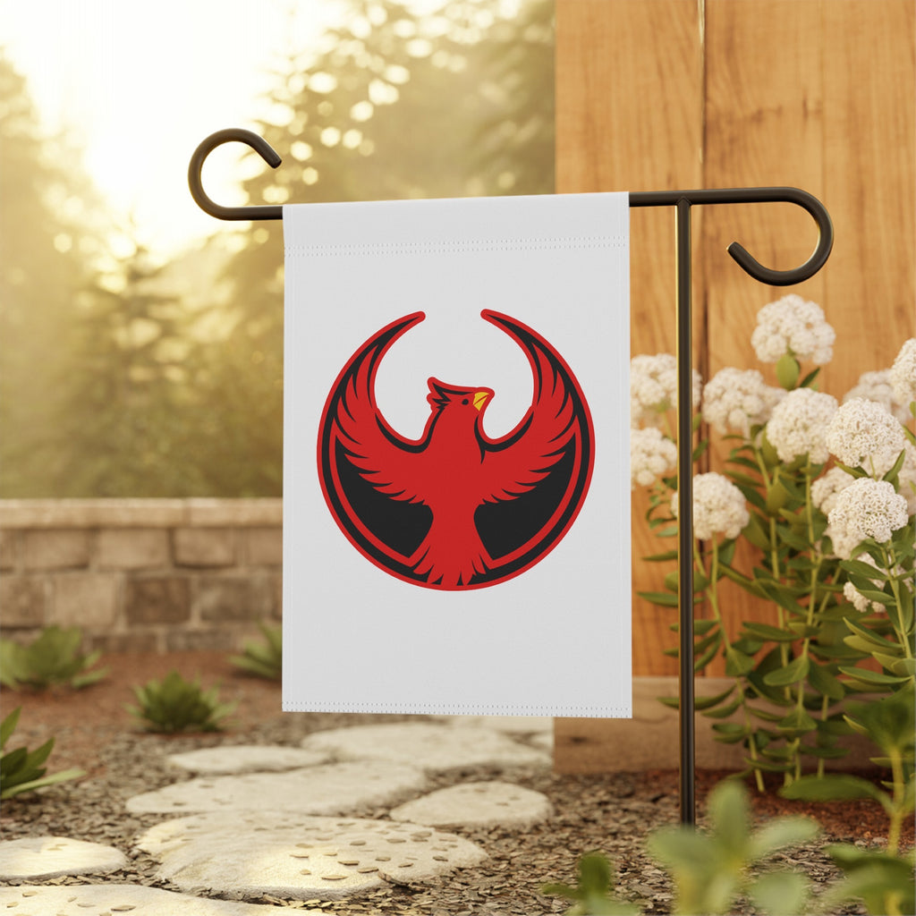 Northern Cardinal Rebel Alliance Garden Flag on a black metal stand in a backyard, featuring a red-and-black phoenix emblem, surrounded by a stone path and flowering shrubs, symbolizing protest and immigrant rights.