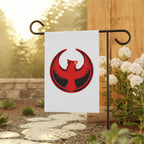 Northern Cardinal Rebel Alliance Garden Flag on a black metal stand in a backyard, featuring a red-and-black phoenix emblem, surrounded by a stone path and flowering shrubs, symbolizing protest and immigrant rights.