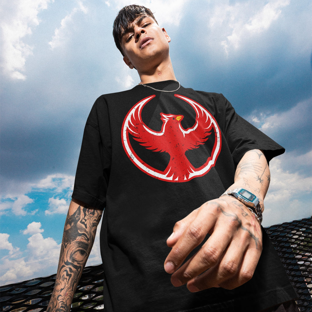 Young man wearing a Northern Cardinal Rebel Alliance T-shirt, featuring a red phoenix graphic on a black tee, embodying themes of protest and community defense, with tattoos and a digital watch visible.