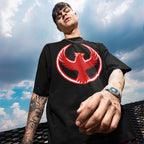 Young man wearing a Northern Cardinal Rebel Alliance T-shirt, featuring a red phoenix graphic on a black tee, embodying themes of protest and community defense, with tattoos and a digital watch visible.