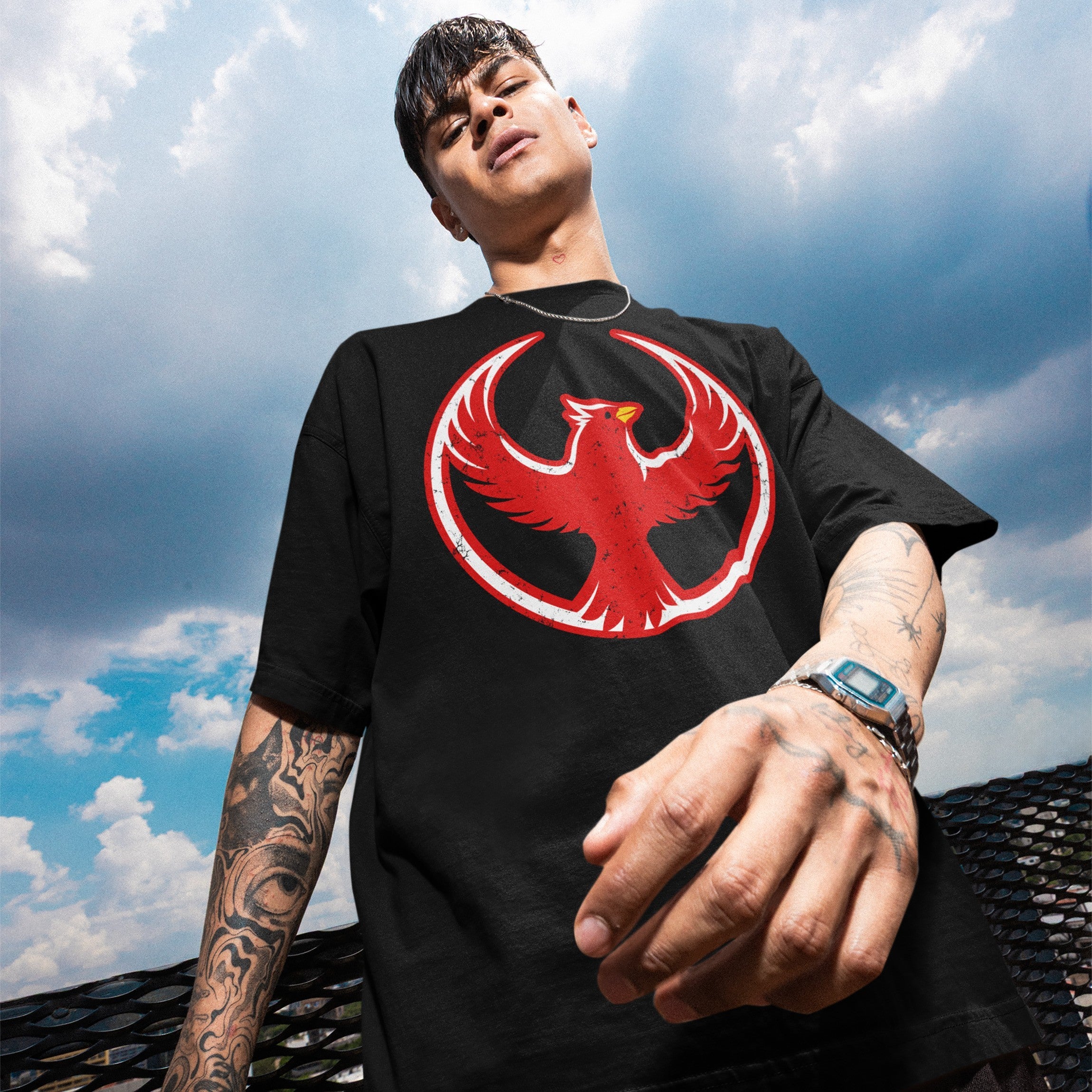 Young man in loose-fitting St. Louis Rebel Alliance T-shirt with a red phoenix emblem, standing against a partly cloudy sky on an elevated platform, embodying activism and equality.
