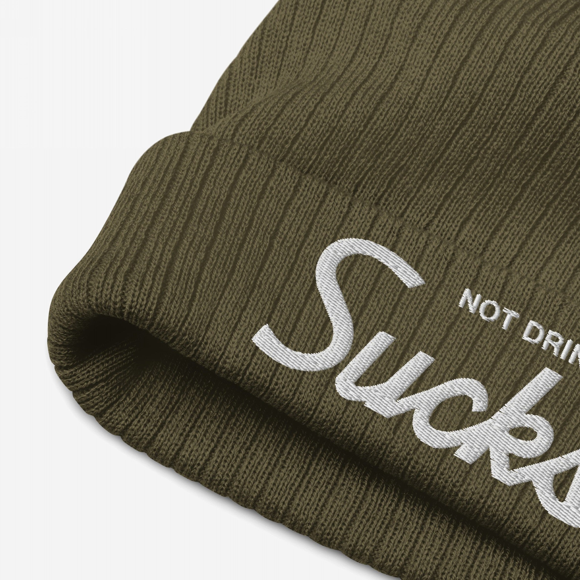 Close-up of the Not Drinking Sucks Hat, an olive-green ribbed knit beanie with a folded cuff, featuring white embroidered text conveying a playful, bold message in retro script.
