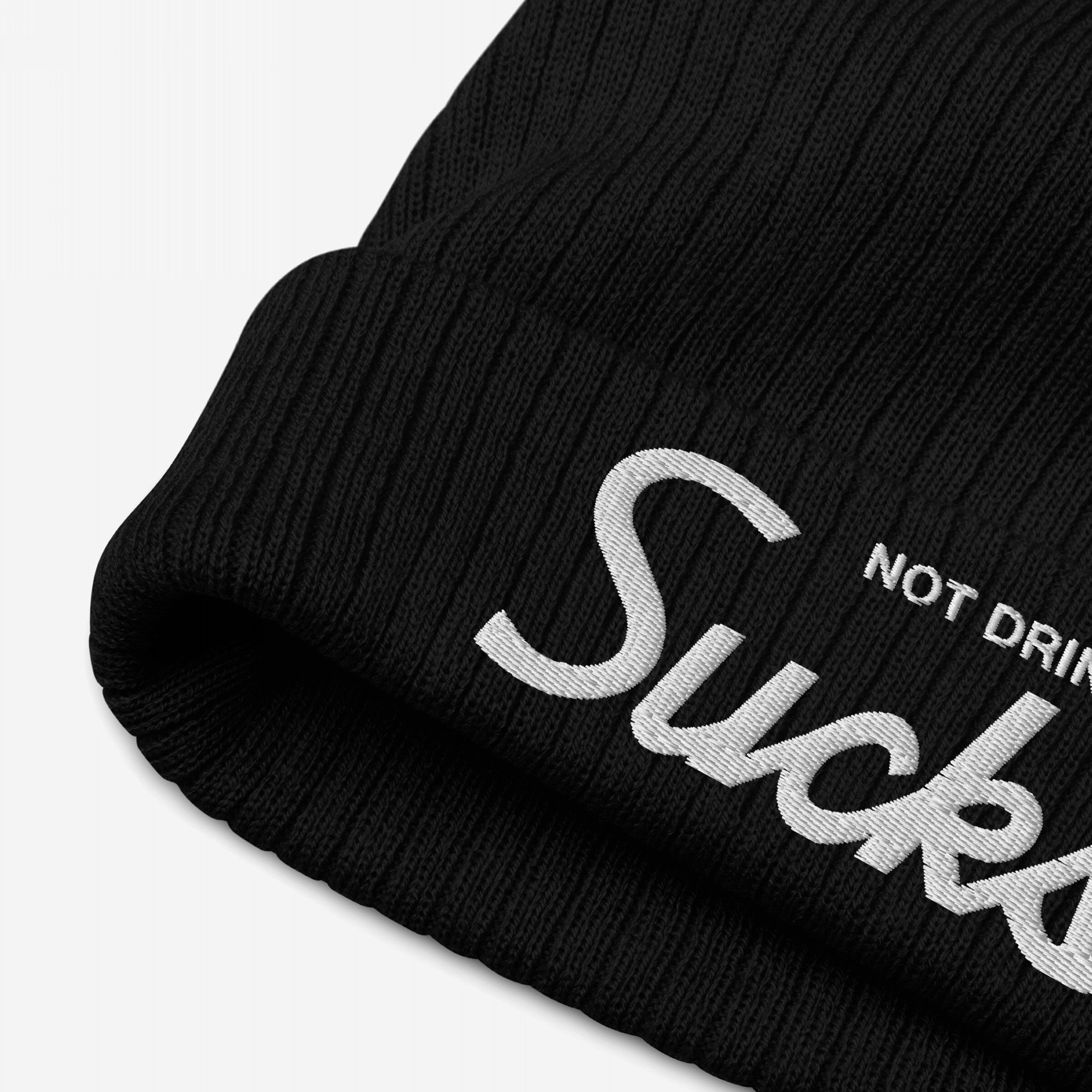 Close-up of the Not Drinking Sucks Hat, a black ribbed knit beanie featuring a fold-over cuff with bold white embroidered script, Sucka, encapsulating a playful, honest party vibe.