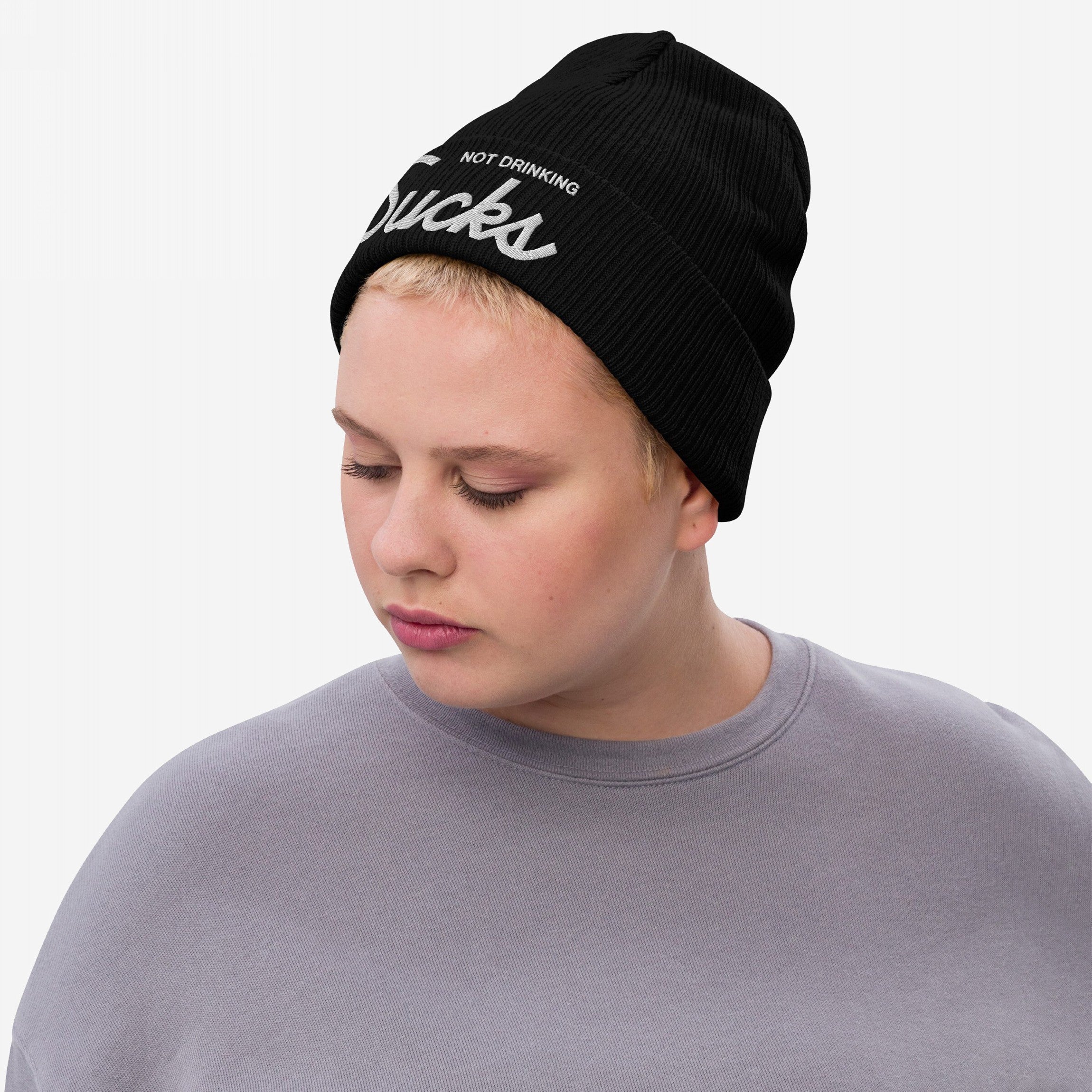 Person wearing the Not Drinking Sucks rib-knit black beanie, showcasing bold white embroidered text. Their light gray top complements the hat's confident, playful message, embodying a lively, inclusive spirit.