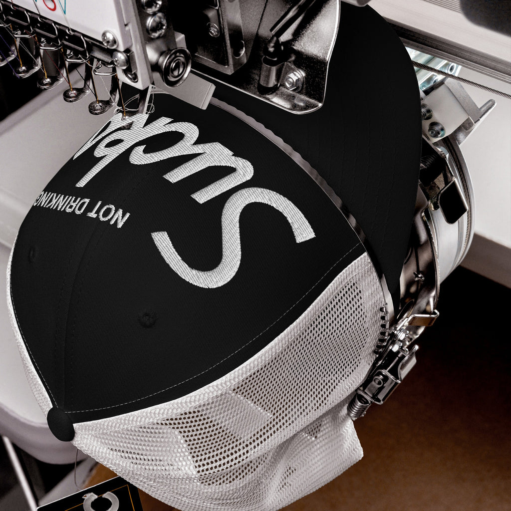 Close-up of an embroidery machine stitching 'Not Drinking Sucks' in white on a black-and-white mesh trucker cap, capturing a bold, playful statement with crisp retro lettering.