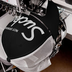 Close-up of an embroidery machine stitching 'Not Drinking Sucks' in white on a black-and-white mesh trucker cap, capturing a bold, playful statement with crisp retro lettering.
