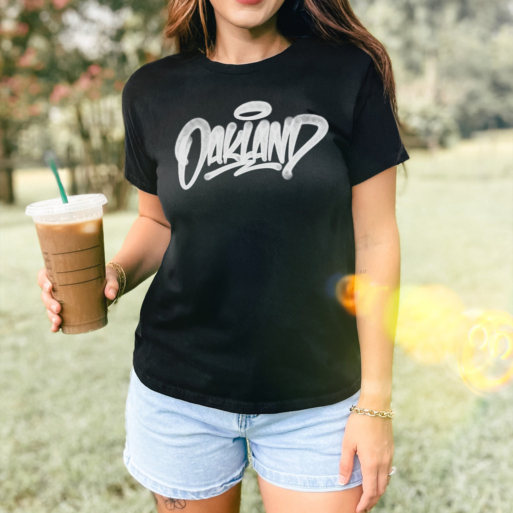 Person wearing the Oakland Handstyle Women's Baby Tee featuring graffiti script, holding iced coffee. Pairing with denim shorts, gold-tone bracelet visible. Background of blurred trees and grass, capturing a casual streetwear vibe.