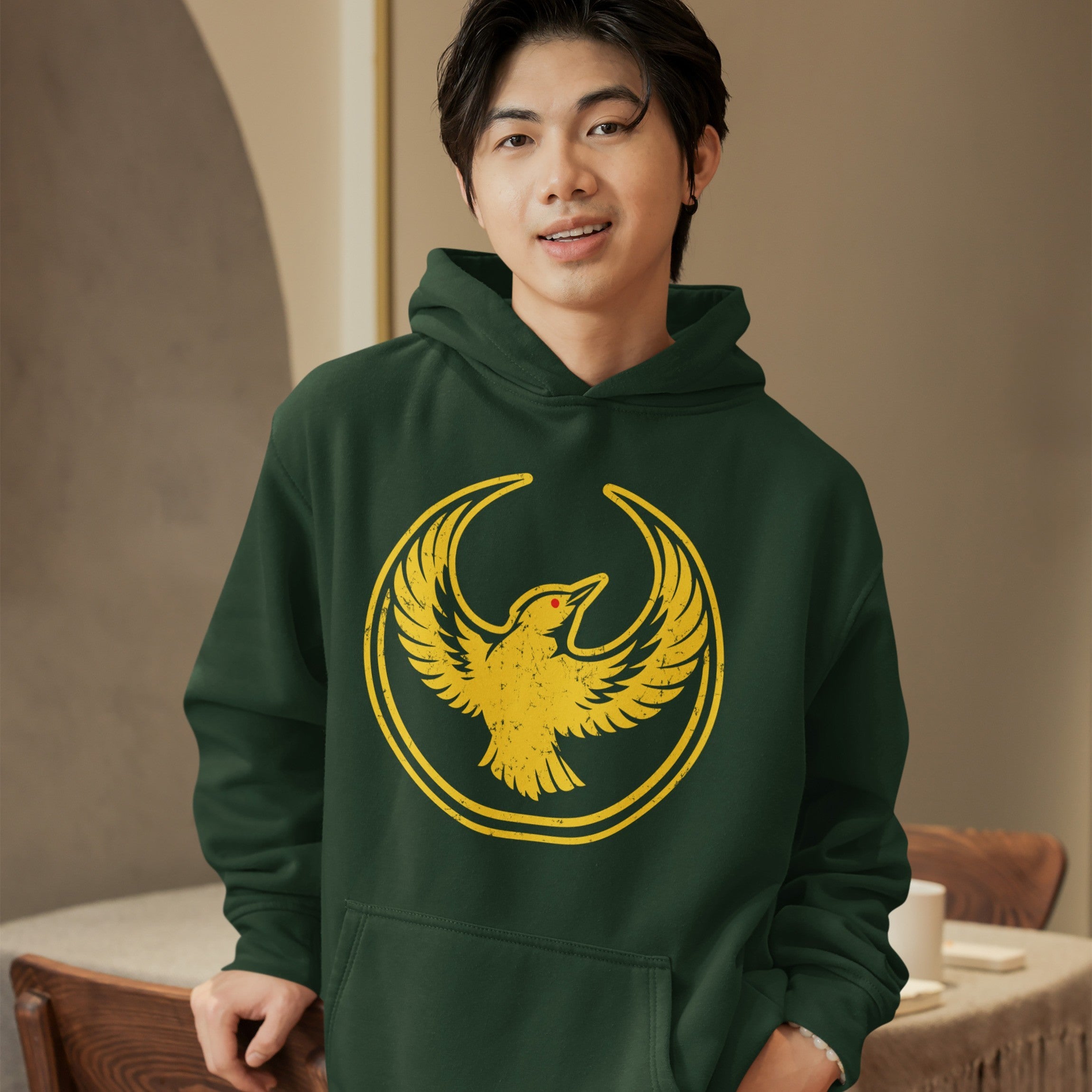 Young person in a forest-green Oregon Rebel Alliance Hoodie, adorned with a golden-yellow bird design, leaning on a wooden chair in a cozy, softly lit room.