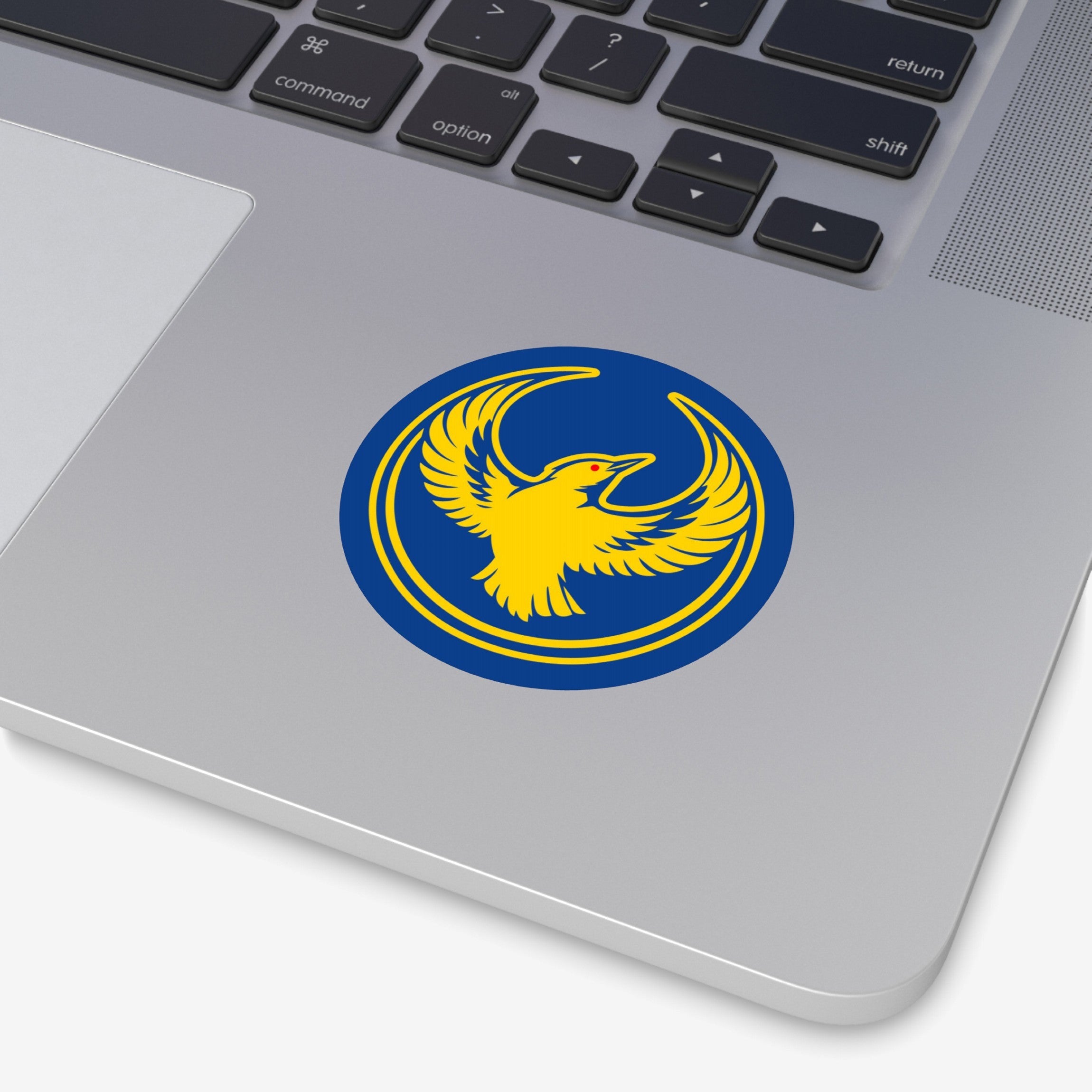 Close-up of a silver laptop with an Oregon Rebel Alliance Sticker on the palm-rest, featuring a stylized yellow phoenix-like bird, symbolizing anti-fascist and abolitionist sentiments.