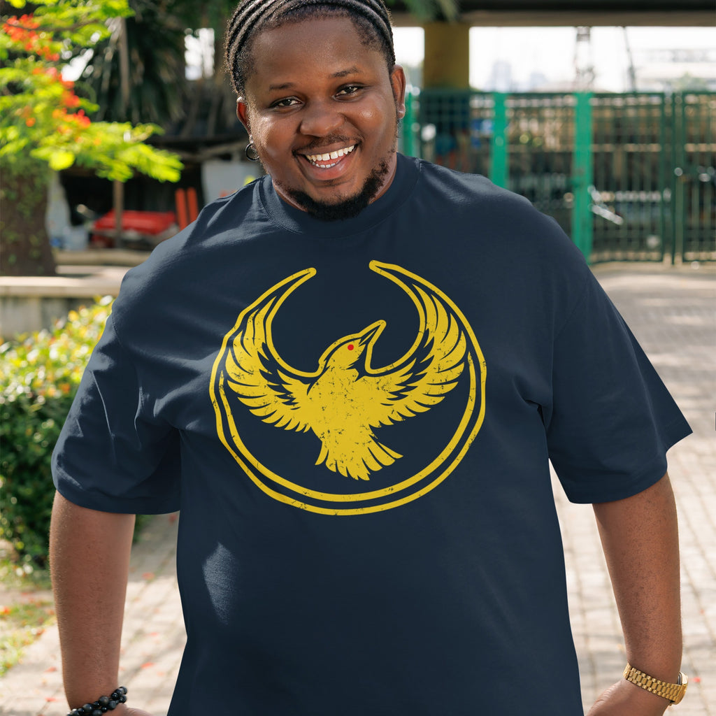 Man wearing an Oregon Rebel Alliance T-shirt featuring a yellow stylized bird emblem, standing outdoors with greenery and a fence in the background, reflecting values of resistance and support for equality.