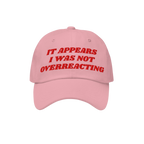 The Overreacting Hat features bold red embroidery on a light pink cap, declaring “IT APPEARS I WAS NOT OVERREACTING,” emb