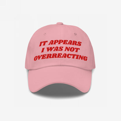 Pink baseball-style cap with red embroidered text, IT APPEARS I WAS NOT OVERREACTING, embodies the Overreacting Hat, designed for vocal progressives opposing political gaslighting. Available in various styles supporting Amnesty International.