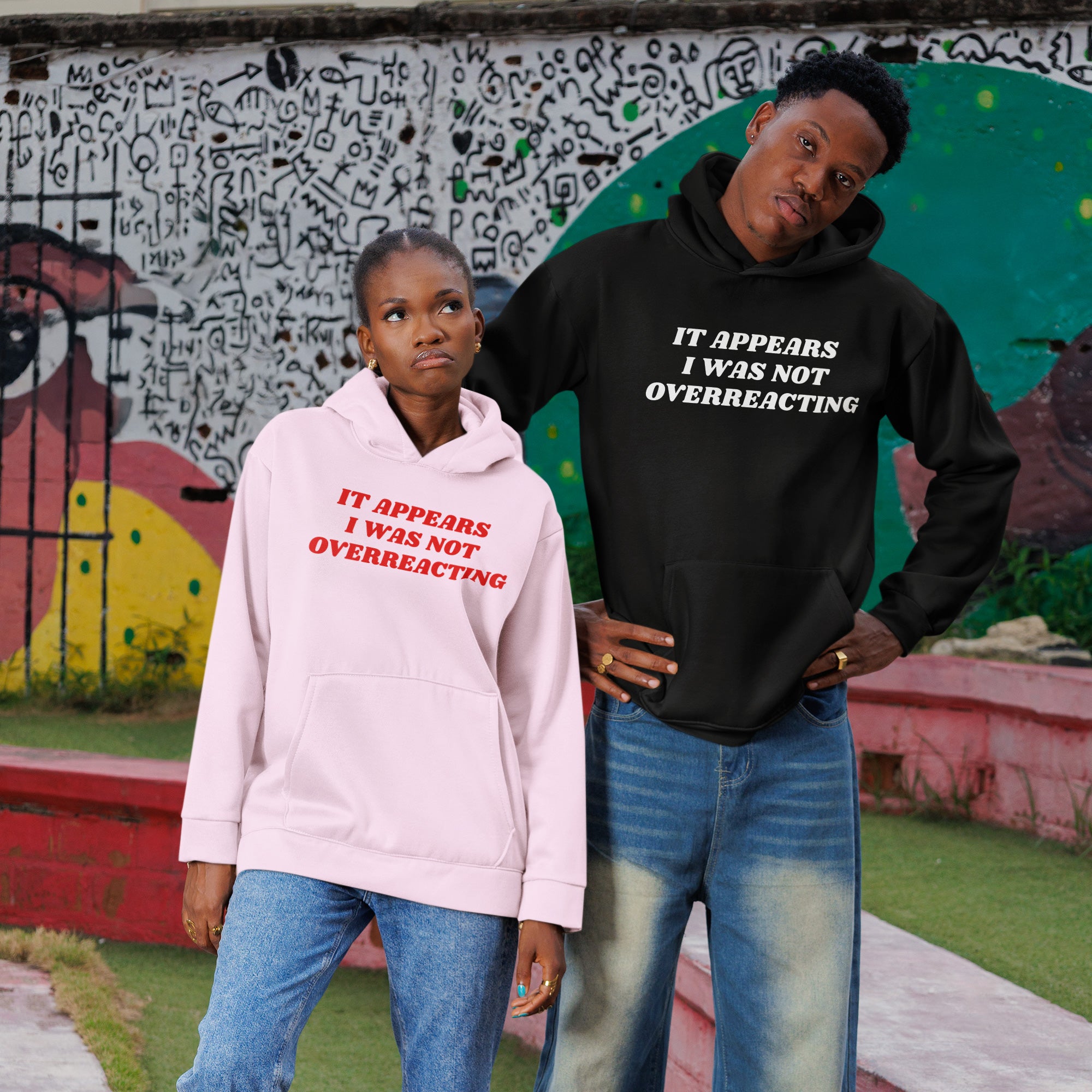 Two young Black models in urban setting, wearing hoodies with Overreacting Shirt slogan. She in pink, he in black, embodying resistance and advocacy through bold, assertive fashion statements.