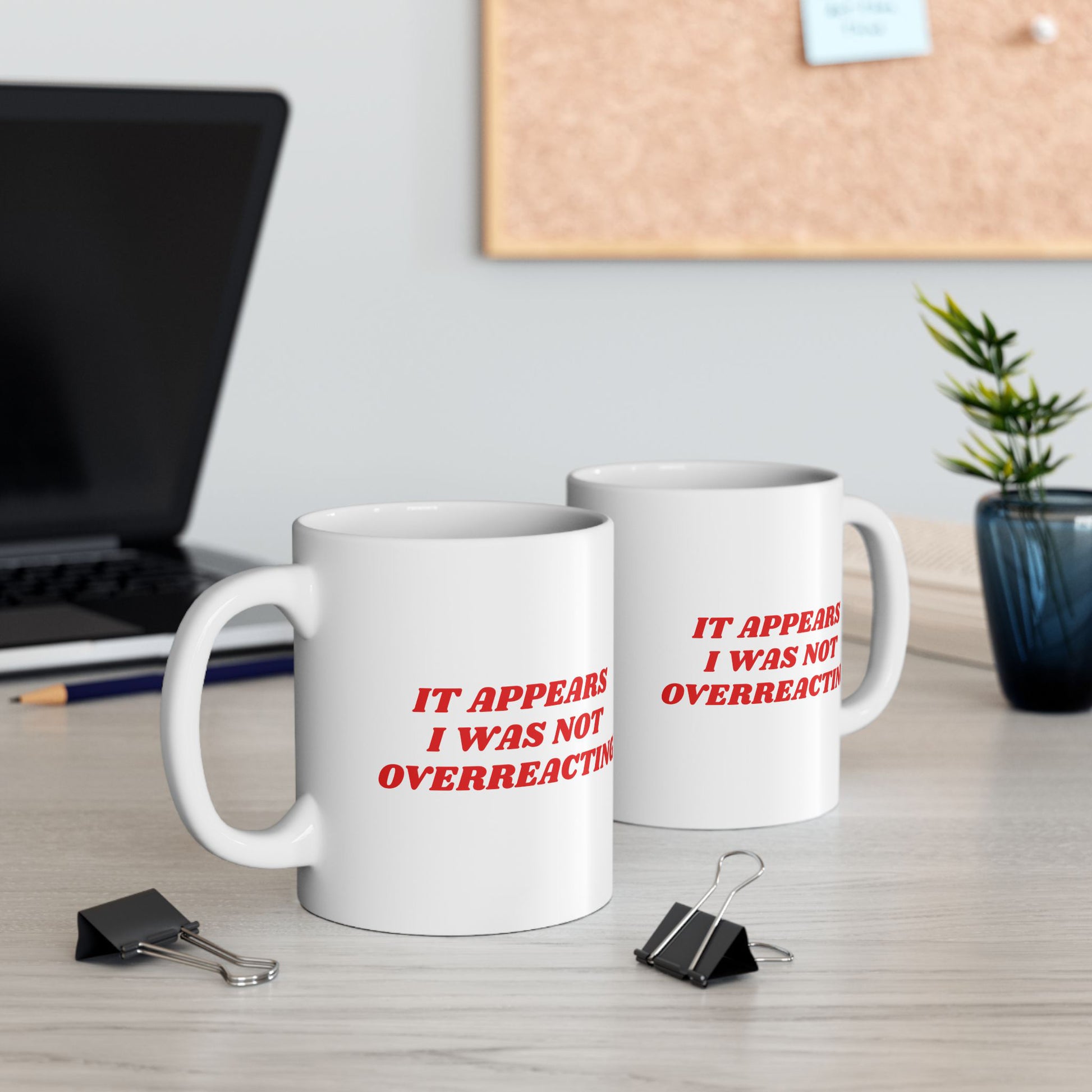 Two white ceramic Overreacting Mugs with red slogan on a desk, surrounded by a laptop, pencil, binder clips, plant, and notebook, embodying bold, progressive statements against fascism.