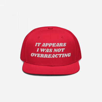 The Overreacting Hat features bold white embroidery reading “IT APPEARS I WAS NOT OVERREACTING” on a flat-brim cap, symbolizing resilience against political gaslighting and supporting activist causes.